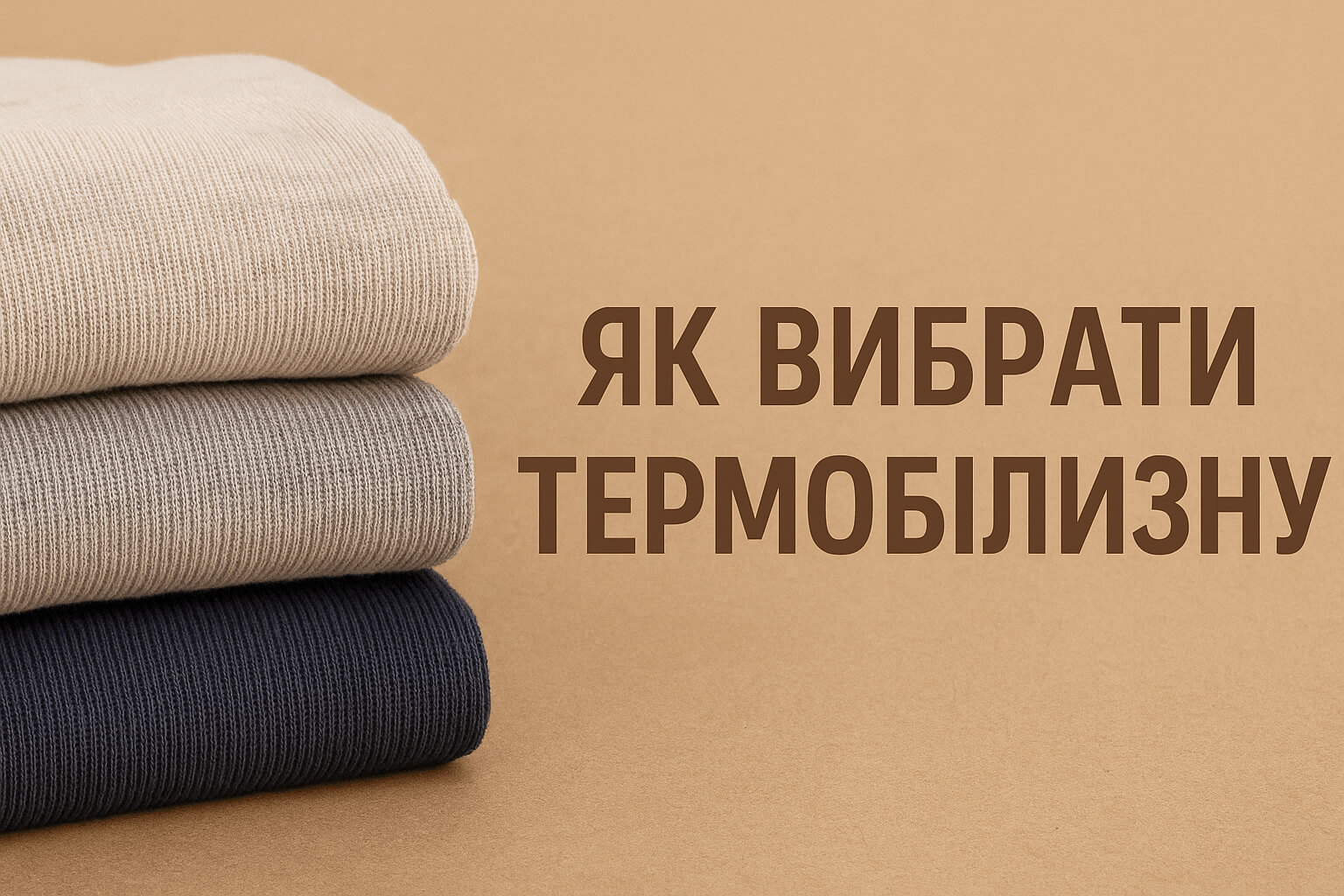 Три складені в стопку термобілизни різних кольорів (бежевий, сірий, темно-синій) на фоні теплого пісочного кольору